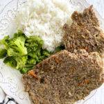 two slices of meatloaf, broccoli and rice on white plate on table next to striped linen